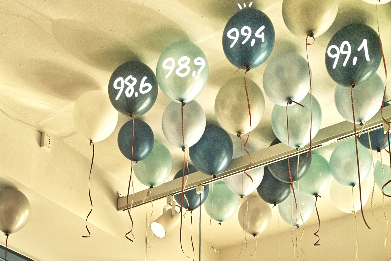 Helium balloons at a ceiling with benchmark scores from 98.6 to 99.4 overlaid, illustrating benchmark saturation in AI models.